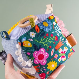 May include: A multi-textured, multi-colored baby toy cube with a wooden ring, fabric loops, and a leather tag. The cube is made of different fabrics, including denim, plaid, and floral prints.