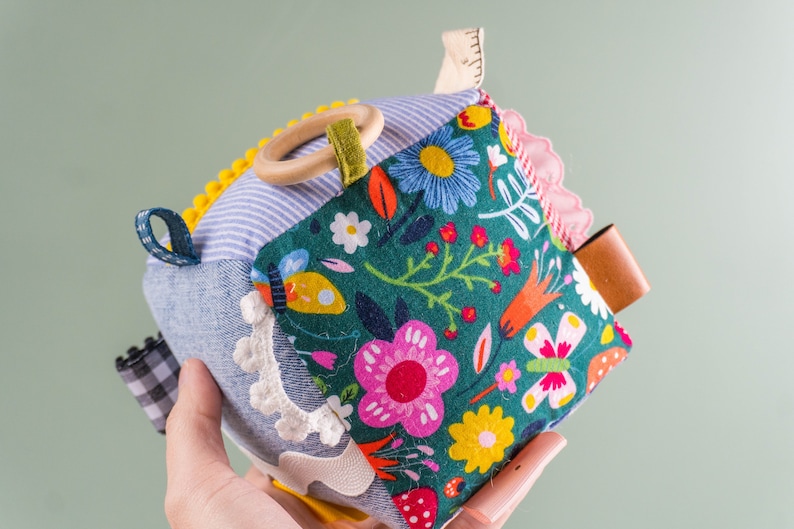May include: A multi-textured, multi-colored baby toy cube with a wooden ring, fabric loops, and a leather tag. The cube is made of different fabrics, including denim, plaid, and floral prints.