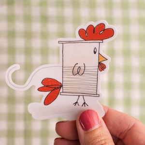 May include: A white and red chicken sticker with a spool of thread for a body. The sticker is on a green and white checkered background.