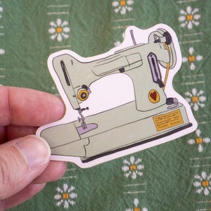 May include: A sticker featuring a vintage-style sewing machine in shades of green, gray, and yellow. The design includes a heart detail and a small label. The sticker is held against a green background with a floral pattern.