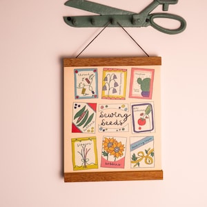 May include: A framed print with a light beige background features colourful illustrations of sewing-related items and seed packets. The text "Sewing Seeds" is displayed in the centre. A vintage green metal scissor-shaped wall hook hangs above.