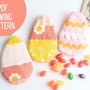 May include: Three fabric Easter egg ornaments, each with a different pattern. The eggs are pink, yellow, and orange with white accents. The eggs are surrounded by colorful jelly beans.