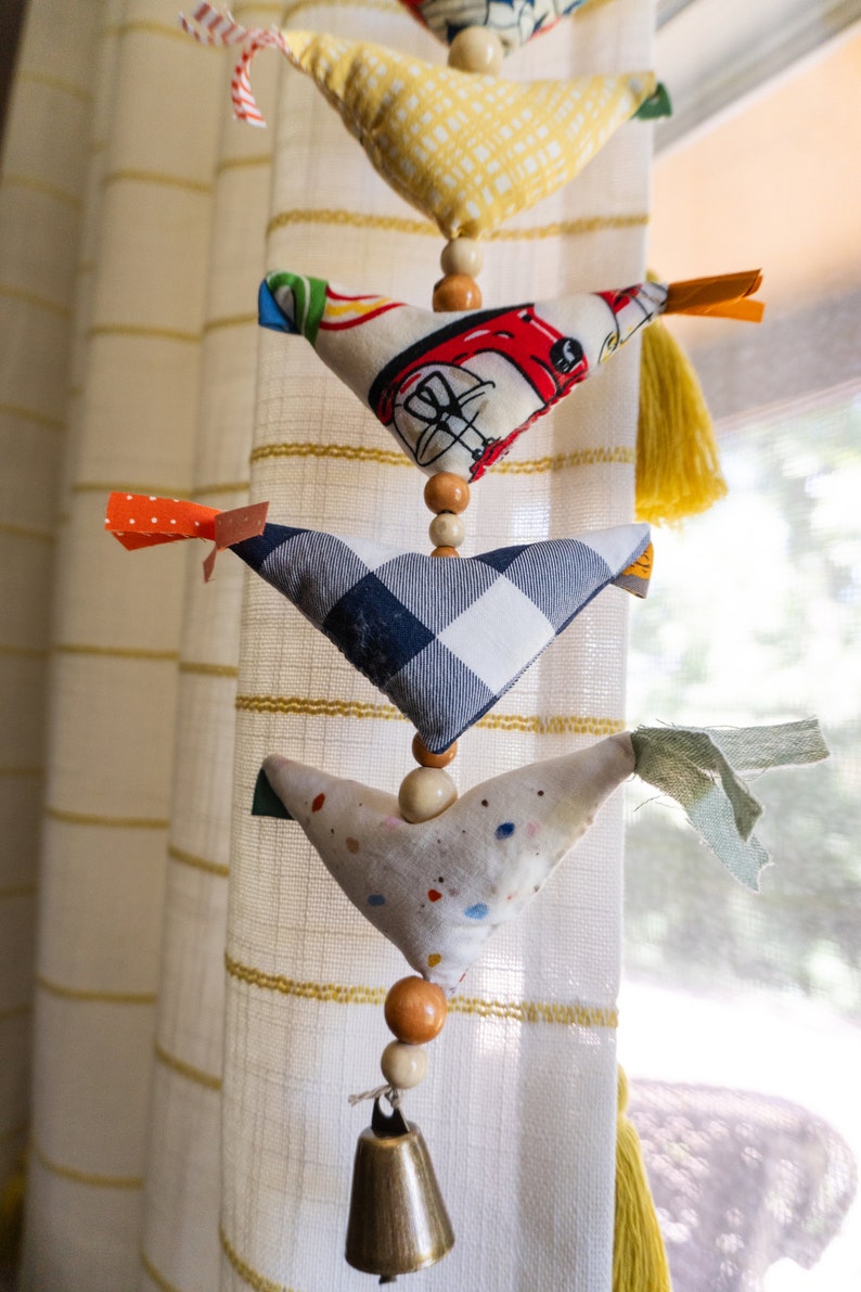 May include: A string of fabric bird ornaments hanging from a white curtain. The birds are made of different fabrics and have different patterns. The string is decorated with wooden beads and a bell at the bottom.