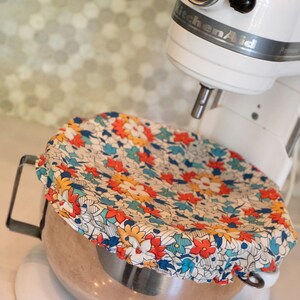 May include: A white KitchenAid stand mixer with a silver bowl and a floral patterned fabric cover. The bowl is filled with a brown mixture.
