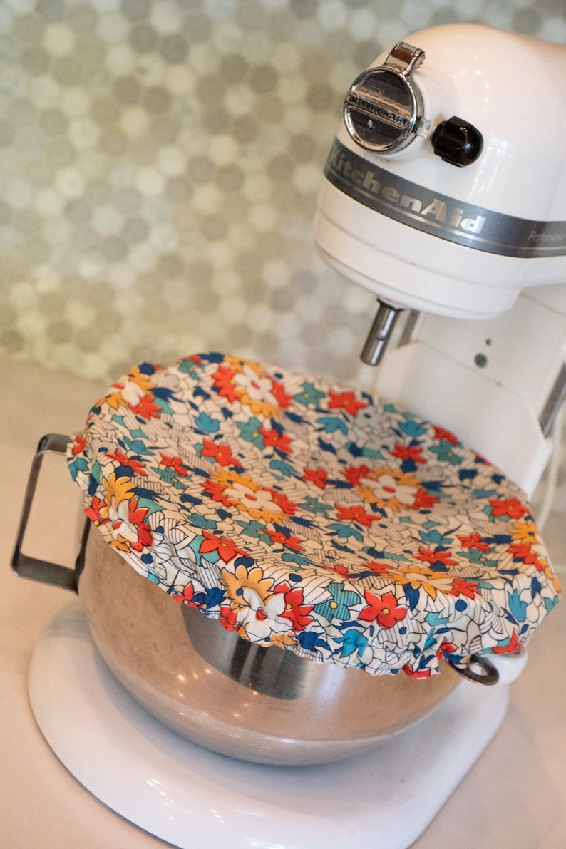 May include: A white KitchenAid stand mixer with a silver bowl and a floral patterned fabric cover. The bowl is filled with a brown mixture.