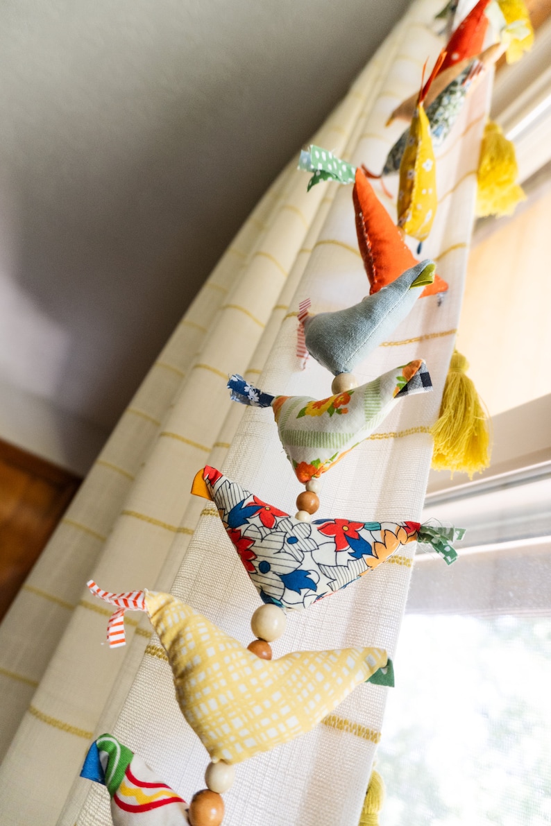 May include: A string of colorful fabric birds hanging from a white curtain. The birds are made of different fabrics and patterns, including floral, polka dot, and geometric designs. The birds are connected by wooden beads.