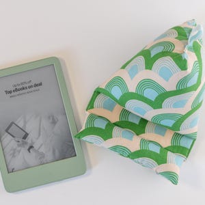 May include: A green and blue patterned pillow with a light beige background. The pillow is folded in half and has a slightly pointed top. The pillow is sitting on a white surface. A green e-reader is also in the image. The e-reader screen displays the text "Up to 50% off Top eBooks on deal Daily Deals & Hottest Picks".