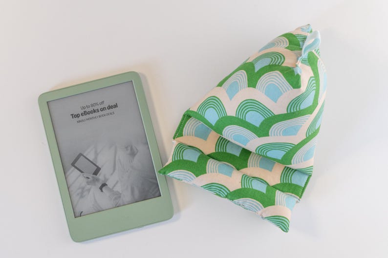 May include: A green and blue patterned pillow with a light beige background. The pillow is folded in half and has a slightly pointed top. The pillow is sitting on a white surface. A green e-reader is also in the image. The e-reader screen displays the text "Up to 50% off Top eBooks on deal Daily Deals & Hottest Picks".