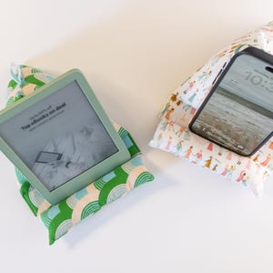 May include: Two fabric phone and tablet stands. One stand is green and white with a rainbow pattern and holds an e-reader with the text "Top e-books on deal" displayed on the screen. The other stand is white with a colorful pattern of people and holds a black smartphone with the time 10:34 displayed on the screen.