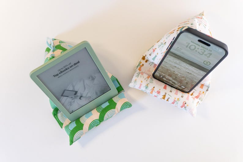 May include: Two fabric phone and tablet stands. One stand is green and white with a rainbow pattern and holds an e-reader with the text "Top e-books on deal" displayed on the screen. The other stand is white with a colorful pattern of people and holds a black smartphone with the time 10:34 displayed on the screen.