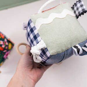 May include: A multi-textured, multi-colored fabric cube toy with a variety of textures, including denim, gingham, and lace. The cube has a white rope tied around it and a white ribbon with a wavy edge.