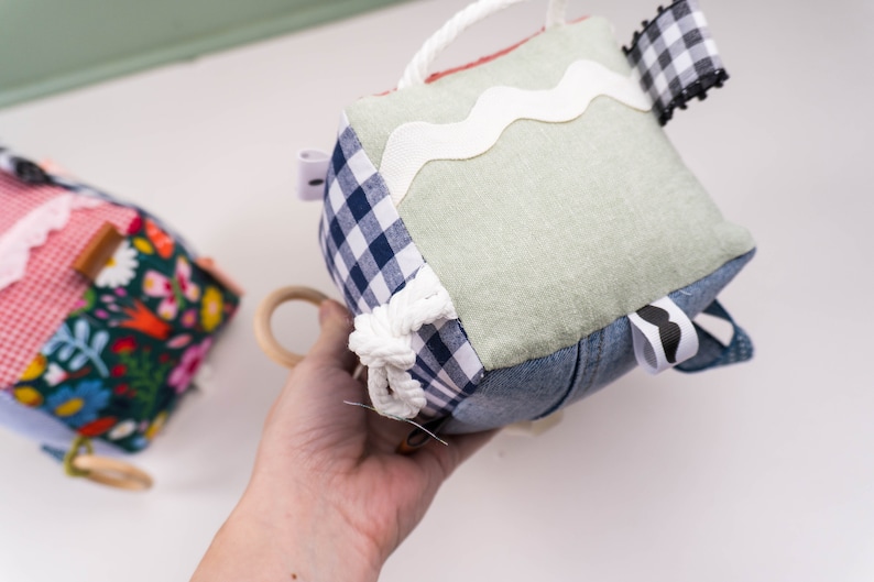 May include: A multi-textured, multi-colored fabric cube toy with a variety of textures, including denim, gingham, and lace. The cube has a white rope tied around it and a white ribbon with a wavy edge.