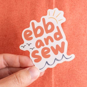 May include: A white sticker with the words "ebb and sew" in orange, featuring a sun, birds, and waves. The sticker is held against an orange background.