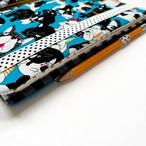 May include: A blue fabric notebook with a black and white dog pattern. The notebook has a white ribbon with black polka dots wrapped around it. A yellow pencil is lying on top of the notebook.