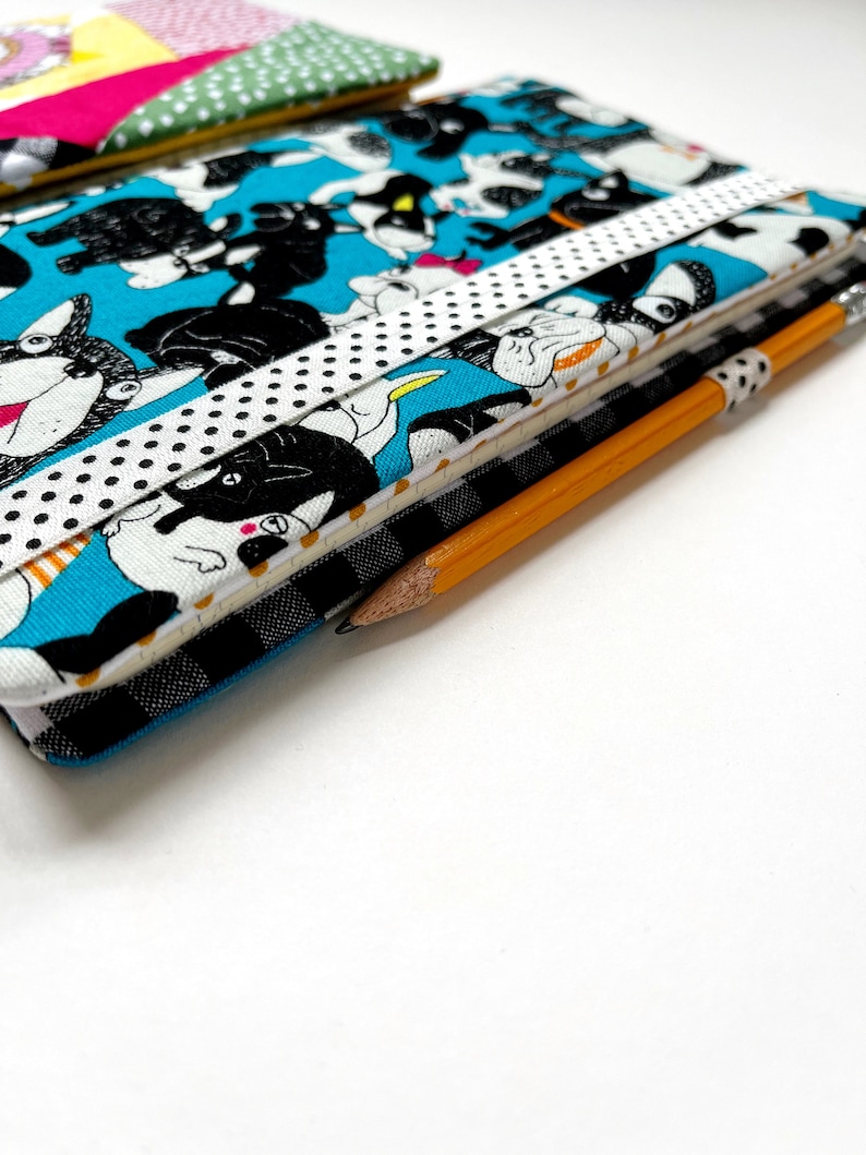 May include: A blue fabric notebook with a black and white dog pattern. The notebook has a white ribbon with black polka dots wrapped around it. A yellow pencil is lying on top of the notebook.