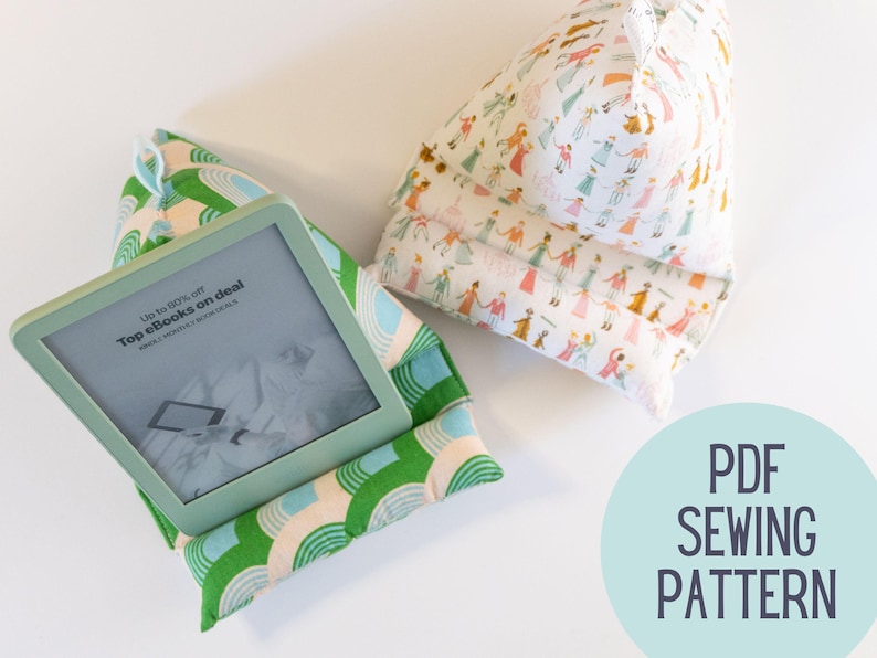 May include: Two green and white fabric book stands with a pattern of rainbows. One stand has a teal e-reader on top with the text "Up to 90% off Top eBooks on deal" on the screen. The other stand has a white fabric with a pattern of colorful people. The text "PDF SEWING PATTERN" is on a teal circle in the bottom right corner.