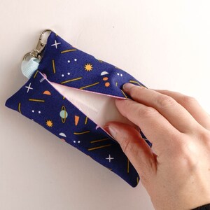 Pode incluir: Uma bolsa de tecido azul com um padr&atilde;o geom&eacute;trico de estrelas, planetas e linhas em amarelo, laranja e branco. A bolsa tem um fecho de correr e um pequeno la&ccedil;o para prender um chaveiro. A bolsa est&aacute; aberta e uma m&atilde;o est&aacute; a alcan&ccedil;ar o interior.