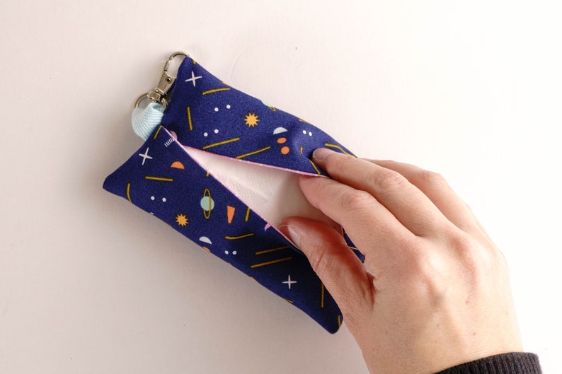 Pode incluir: Uma bolsa de tecido azul com um padr&atilde;o geom&eacute;trico de estrelas, planetas e linhas em amarelo, laranja e branco. A bolsa tem um fecho de correr e um pequeno la&ccedil;o para prender um chaveiro. A bolsa est&aacute; aberta e uma m&atilde;o est&aacute; a alcan&ccedil;ar o interior.