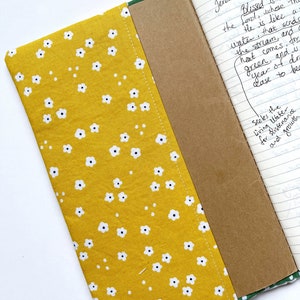 May include: A yellow fabric notebook cover with a white floral pattern. The cover is open to reveal a brown notebook with lined pages. The notebook has handwritten text on the pages, including the Bible verse Jeremiah 17:7+8.