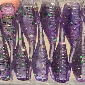May include: A collection of translucent purple soft plastic fishing lures featuring a ribbed body design and embedded green and gold glitter flakes. Each lure has a tapered tail section and a segmented appearance, arranged in a parallel row.