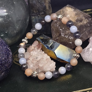 May include: A beaded bracelet featuring a mix of light blue, peach, and dark gray gemstones, interspersed with silver-colored metal beads. The bracelet is displayed on a dark green surface with other crystals and stones in the background.