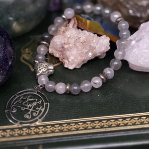 May include: A grey beaded bracelet with a silver charm featuring a detailed symbol and a leopard head. The bracelet is displayed with a cluster of crystals on a dark green surface.