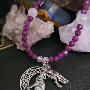 May include: A purple beaded necklace with a silver pendant featuring a howling wolf and a rabbit. The necklace includes white and clear beads. The background includes crystals and a dark green surface.