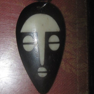 May include: A black and white wooden pendant in the shape of a face. The face has a simple design with two eyes, a nose, and a mouth.
