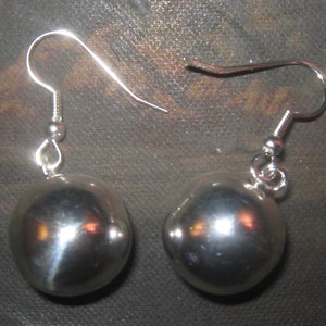May include: A pair of silver-colored ball earrings. Each earring features a large, smooth, spherical bead suspended from a simple hook. The earrings reflect light, creating highlights on their surface. The hooks are a simple, curved design.