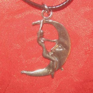 May include: A silver-toned pendant necklace featuring a crescent moon design with a figure embracing the moon. The pendant hangs from a black cord. The background is a textured red fabric.