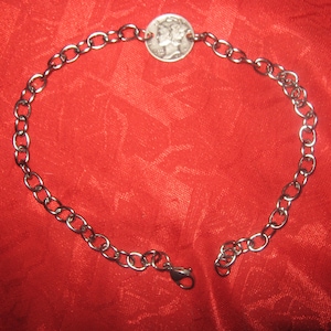 May include: A silver-colored chain bracelet with a coin pendant. The bracelet features a series of oval links and a lobster clasp closure. The coin in the center has a detailed design. The bracelet is displayed on a red background.