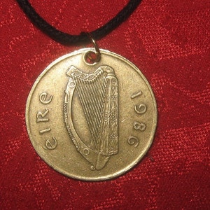 K&ouml;nnte beinhalten: Ein goldfarbener irischer M&uuml;nzenanh&auml;nger mit der Aufschrift "EIRE 1986" und einem Harfendesign. Die M&uuml;nze ist an einer schwarzen Schnur befestigt.