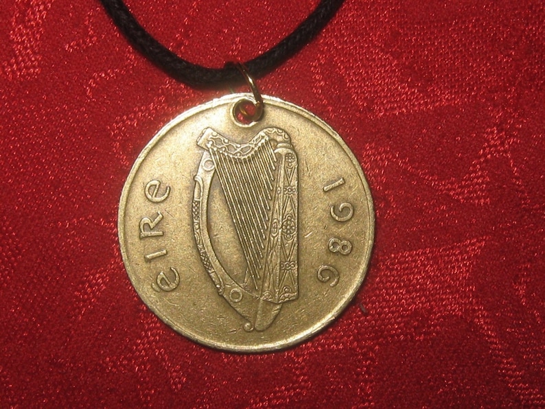 K&ouml;nnte beinhalten: Ein goldfarbener irischer M&uuml;nzenanh&auml;nger mit der Aufschrift "EIRE 1986" und einem Harfendesign. Die M&uuml;nze ist an einer schwarzen Schnur befestigt.