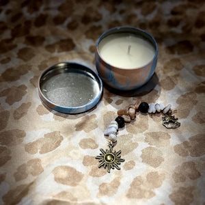 May include: A small, blue and pink candle in a tin container with a white wax interior. The candle is sitting on a brown and white patterned fabric with a gold sun charm on a beaded chain.