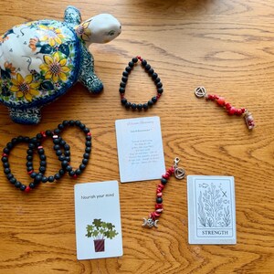 May include: A ceramic turtle figurine with a sunflower pattern, four black lava bead bracelets with red accents, two beaded charms with silver-tone symbols, and two cards. One card features a plant illustration and the text "Nourish your mind," while the other displays a coral drawing and the word "STRENGTH."