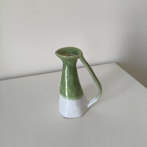 Puede incluir: Un pequeño jarrón de cerámica verde y blanco con asa. El jarrón está esmaltado en color verde claro en la mitad superior y blanco en la mitad inferior.