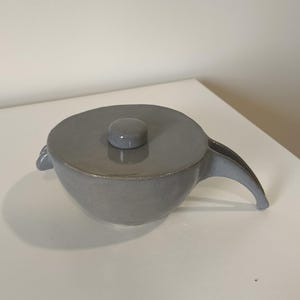 Puede incluir: Una tetera de cerámica gris con tapa y una sola asa. La tetera tiene un cuerpo liso y redondeado y una pequeña perilla en la tapa.