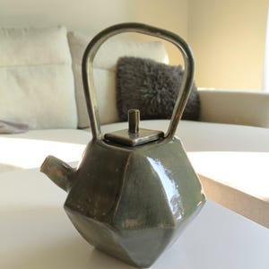 May include: A faceted, olive-green ceramic teapot with a curved handle and a square lid. The teapot has a spout and sits on a white surface. The background includes a couch and a decorative pillow.