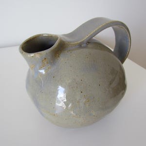 Puede incluir: Una jarra de cerámica gris con un esmalte moteado y un cuerpo redondeado. La jarra tiene un asa que está unida al lado de la jarra.