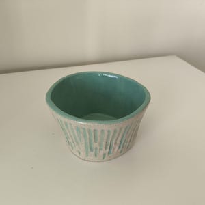 Puede incluir: Un pequeño cuenco de cerámica verde claro con líneas verticales blancas. El cuenco está esmaltado por dentro y tiene una forma ligeramente redondeada.