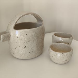 Peut inclure: Un pichet en céramique blanche avec une anse et deux tasses assorties. Le pichet et les tasses ont une finition mouchetée.