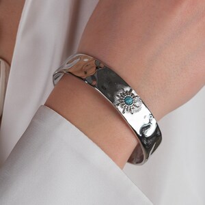 May include: A silver hammered bangle bracelet with a turquoise stone and silver accents. The bracelet is on a person's wrist.