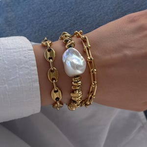 Peut inclure: Ensemble de bijoux dorés comprenant un bracelet chaîne, un bracelet avec une grosse perle et une bague à plusieurs anneaux. Les bijoux sont portés avec une chemise blanche et un jean bleu. Les bracelets sont de styles différents.