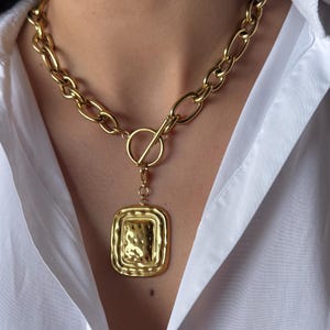 Puede incluir: Un collar dorado con una cadena gruesa y un colgante cuadrado. El colgante tiene una superficie texturizada y cuelga de un cierre de palanca. El collar se lleva sobre una camisa blanca.