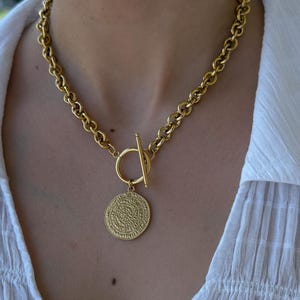 May include: Gold-coloured necklace with a chunky chain and a circular pendant with an intricate design. The necklace has a toggle clasp. The pendant hangs from a small ring. The necklace is worn against a white blouse.