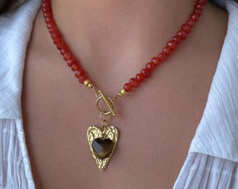 Pode incluir: Um colar de contas vermelhas com fecho dourado e um pendente em forma de coração. O pendente apresenta uma moldura dourada e um coração de pedra castanha. O colar é usado sobre uma camisa branca.