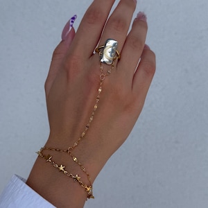 Peut inclure: Un bracelet de main doré avec des étoiles et une bague avec une face rectangulaire ornée d'un croissant de lune. Le bijou est porté sur une main aux ongles longs et manucurés, sur fond blanc.