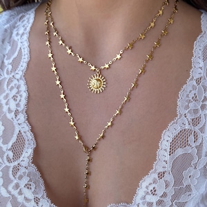 Puede incluir: Un collar dorado en capas con un colgante de sol y abalorios en forma de estrella. El collar se lleva sobre una camiseta sin mangas blanca con detalles de encaje. El colgante de sol está centrado en el pecho.