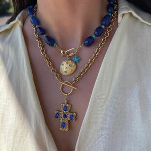 Può includere: Una collana a strati con una catena dorata e fili di perline blu. La collana include un ciondolo dorato con un design a conchiglia e un ciondolo a forma di stella marina blu. Un ciondolo a croce dorata con pietre preziose blu è appeso sotto.