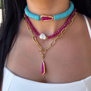 May include: A layered necklace set featuring a top choker of turquoise disc beads with a gold-rimmed magenta pendant, a middle strand of small magenta beads with a white baroque pearl, and a bottom gold chain with a magenta teardrop pendant.